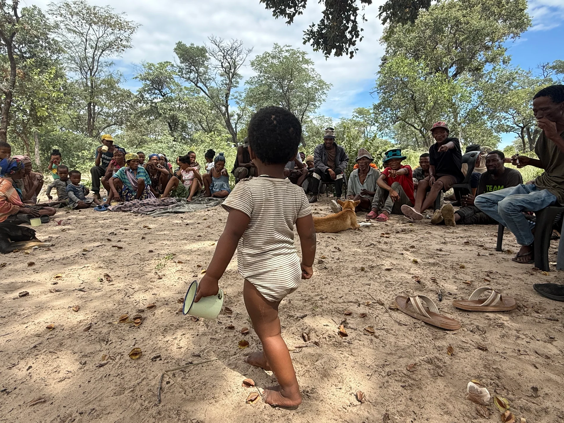 A child holding a cup walks towards a community gathering beneath the trees